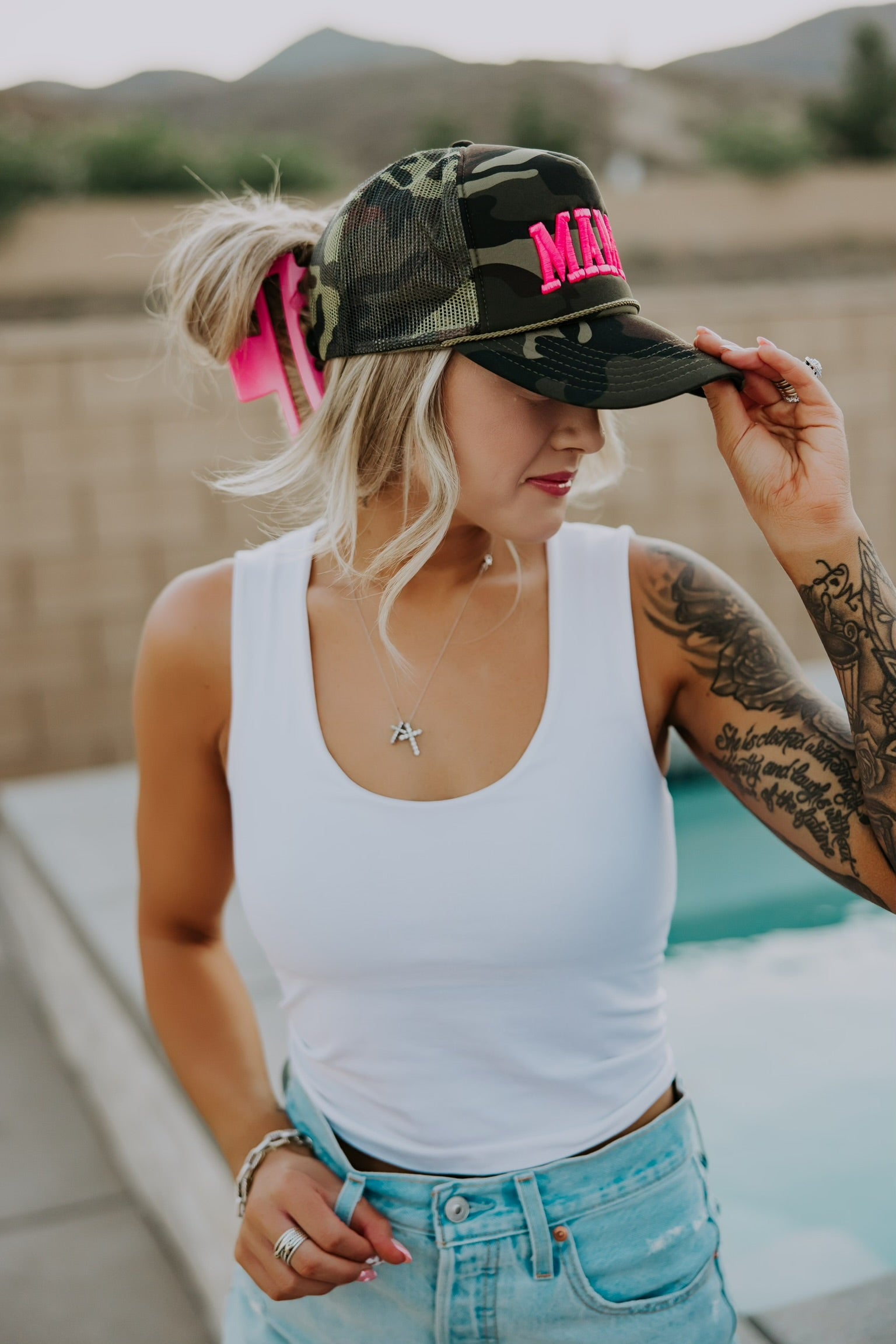A woman with thick hair in a ponytail, secured by the Pink Claw Clip - XL from Mama X™ Brand, stands poolside in a white tank and denim shorts, holding a camouflage "MAMA" cap. She displays her tattooed arm and silver necklace.
