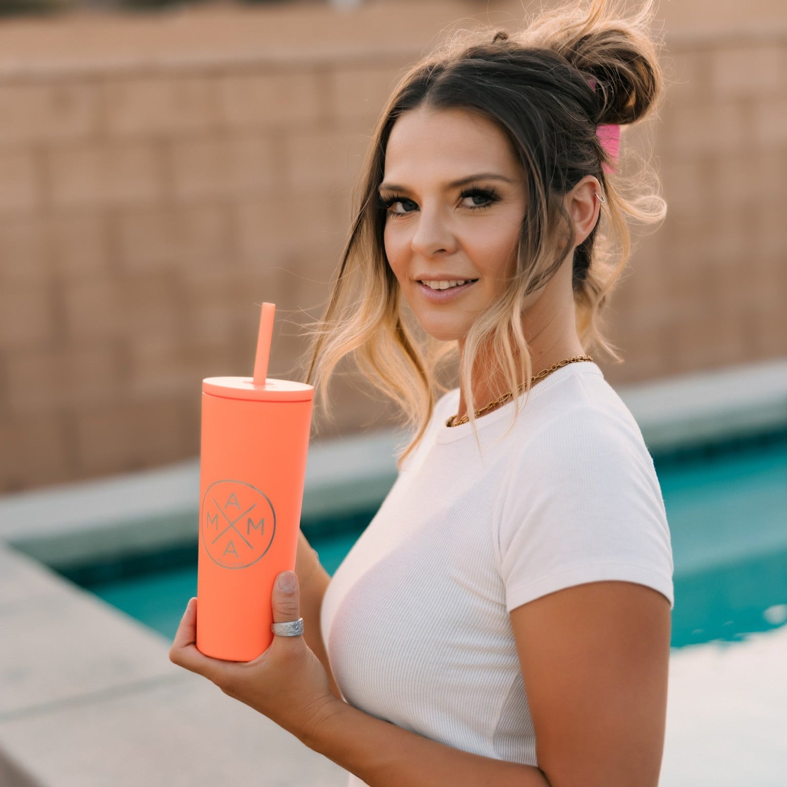 A woman with shoulder-length hair in a half-up style, wearing a white top and denim shorts, sits by the pool smiling at the camera while holding the Mama X™ Brand Coral Tumbler with a straw.