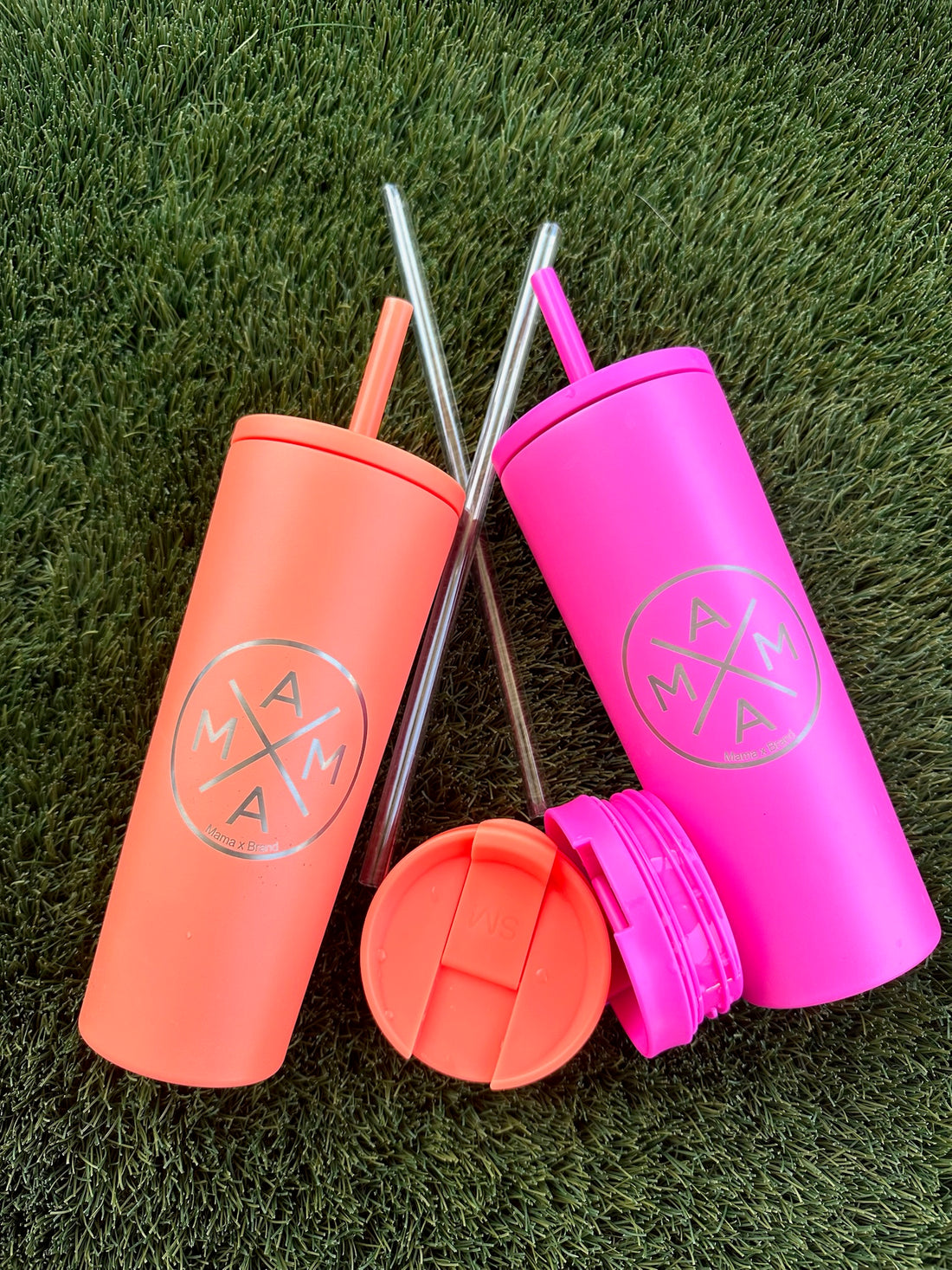 Two reusable Mama X™ Brand tumblers—one hot pink, one orange—featuring a circular "MAMA" logo rest on green grass. Each comes with a metal straw and the lids are placed in front of the tumblers.