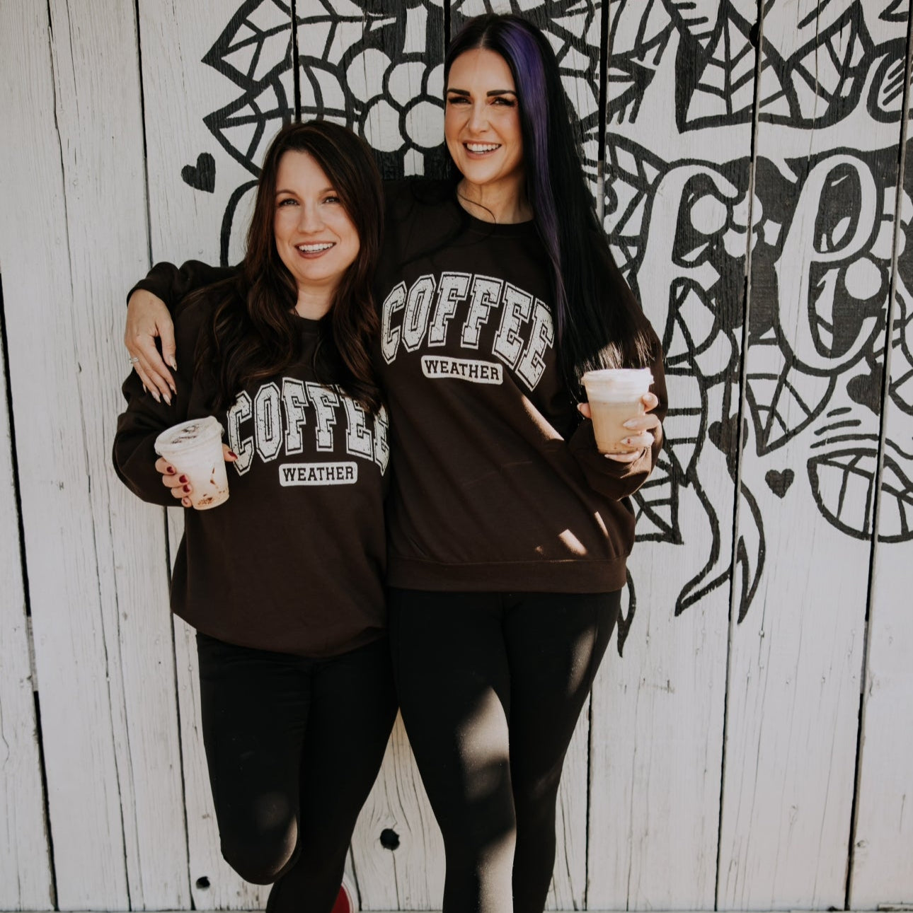 Two women stand arm-in-arm, smiling in front of a white wall with black floral art, both wearing matching Mama X™ Brand Coffee Weather Crewnecks and black leggings, holding iced coffees—the perfect look for sweater weather.