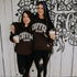Two women stand arm-in-arm, smiling in front of a white wall with black floral art, both wearing matching Mama X™ Brand Coffee Weather Crewnecks and black leggings, holding iced coffees—the perfect look for sweater weather.