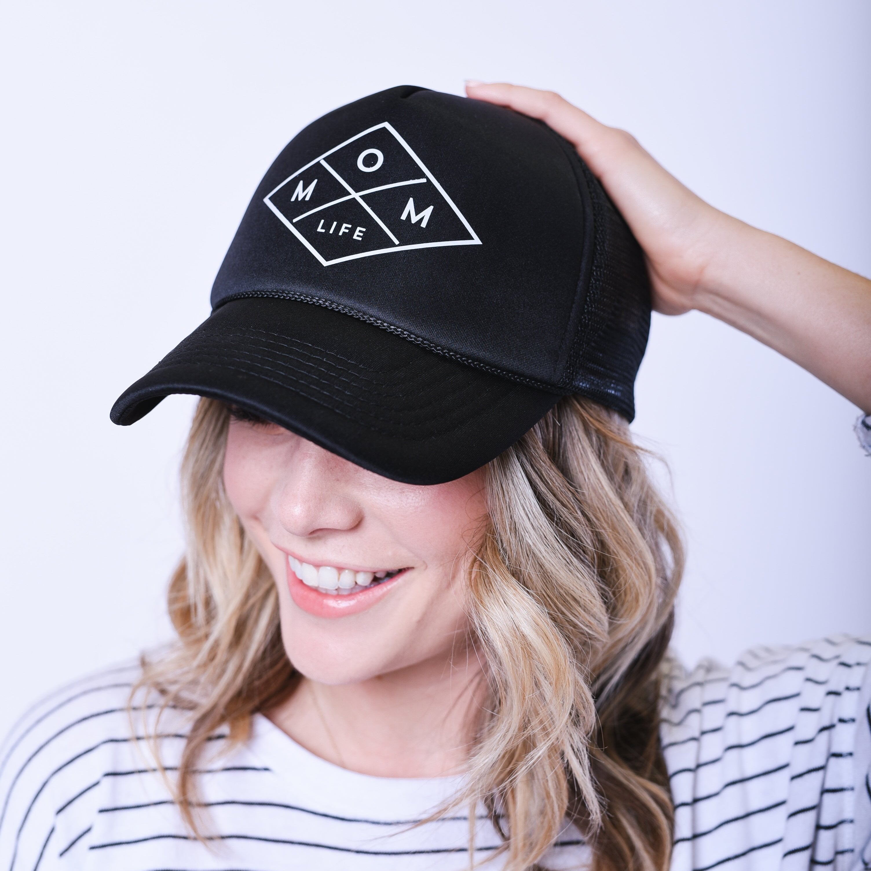A blonde woman, dressed in a white and black striped shirt, smiles while wearing the high-quality "Mom Life Foam Hat - Black" from Mama X™ Brand. Holding the back of her head with her right hand, she stands against a light, plain background that highlights her relaxed vibe.