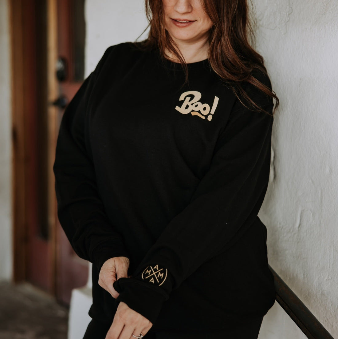 A woman with long brown hair adjusts her sleeve while wearing the Mama X™ Brand Boo Crewneck sweatshirt indoors, standing in front of a white textured wall and wooden door.