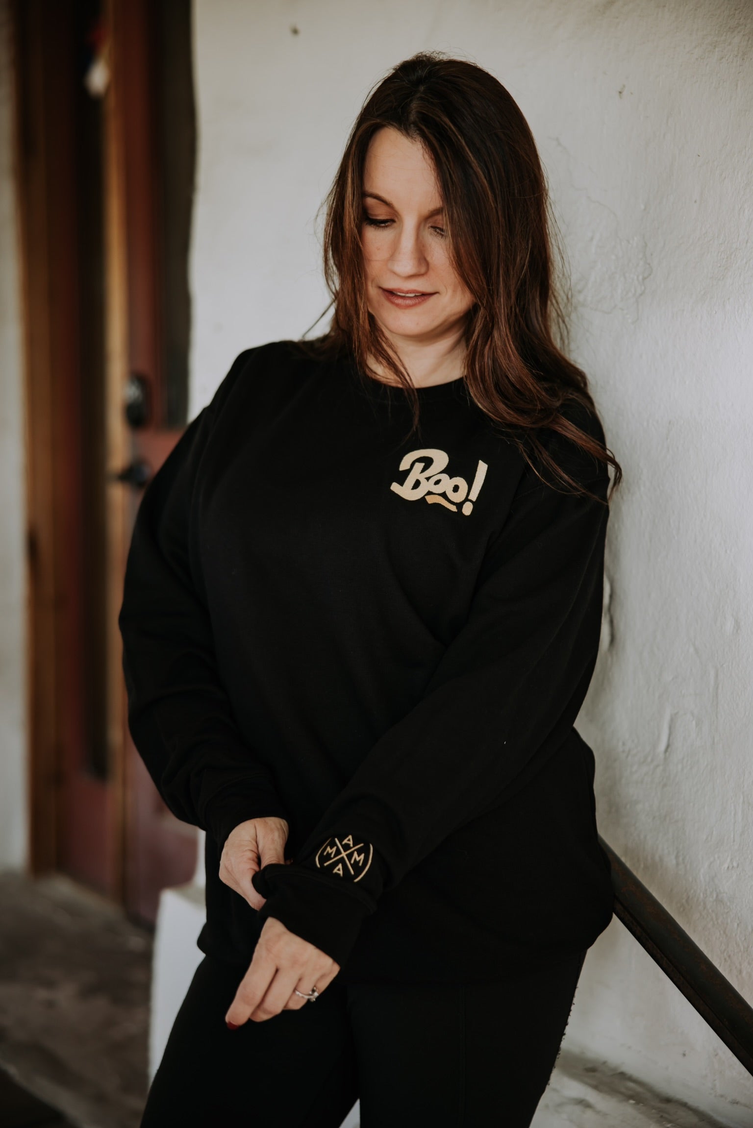 A woman with long brown hair adjusts her sleeve while wearing the Mama X™ Brand Boo Crewneck sweatshirt indoors, standing in front of a white textured wall and wooden door.