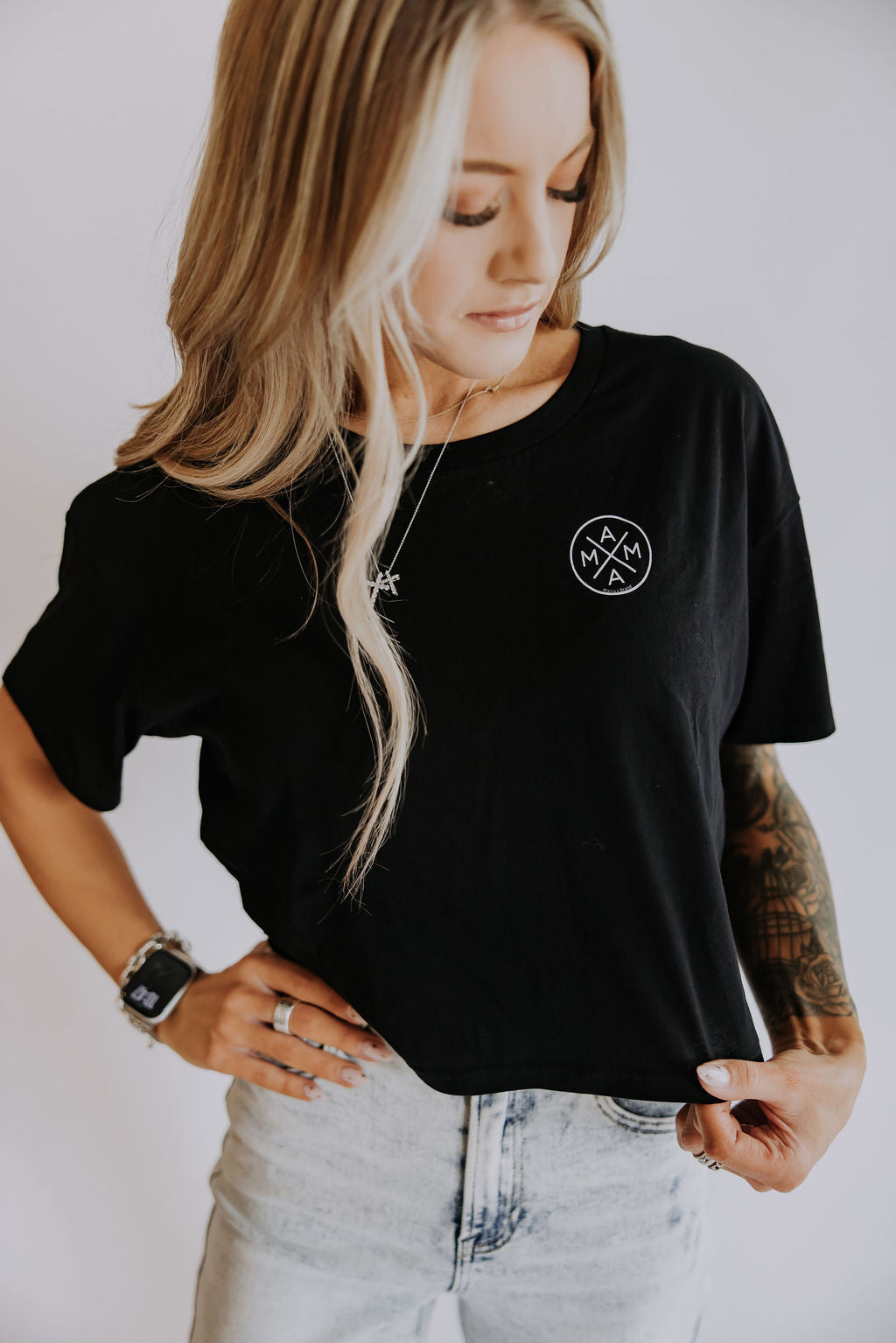 A woman with long blonde hair wears the Mama X™ Brand Mama X crop top, featuring a small white circular logo, paired with light high-waisted jeans. She looks down, holding the hem to reveal her tattooed arm, watch, and rings.