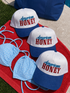 Three Mama X™ Brand "American Honey" white and blue baseball caps with red and blue text are on a red towel by the pool, next to a blue polka-dot bikini and a blue bag.