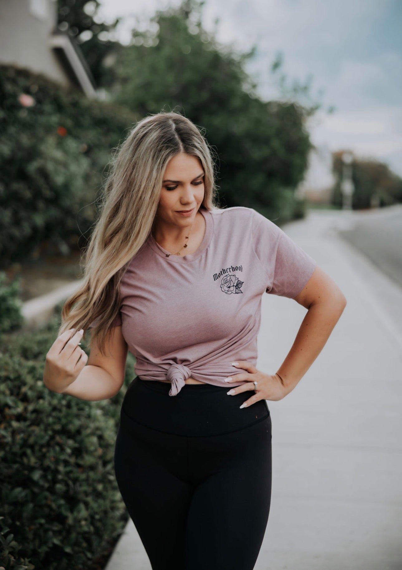 A woman with long, wavy hair stands on a sidewalk beside greenery, wearing a mauve Motherhood Tshirt from Mama X™ Brand and black leggings, looking down with one hand touching her hair.
