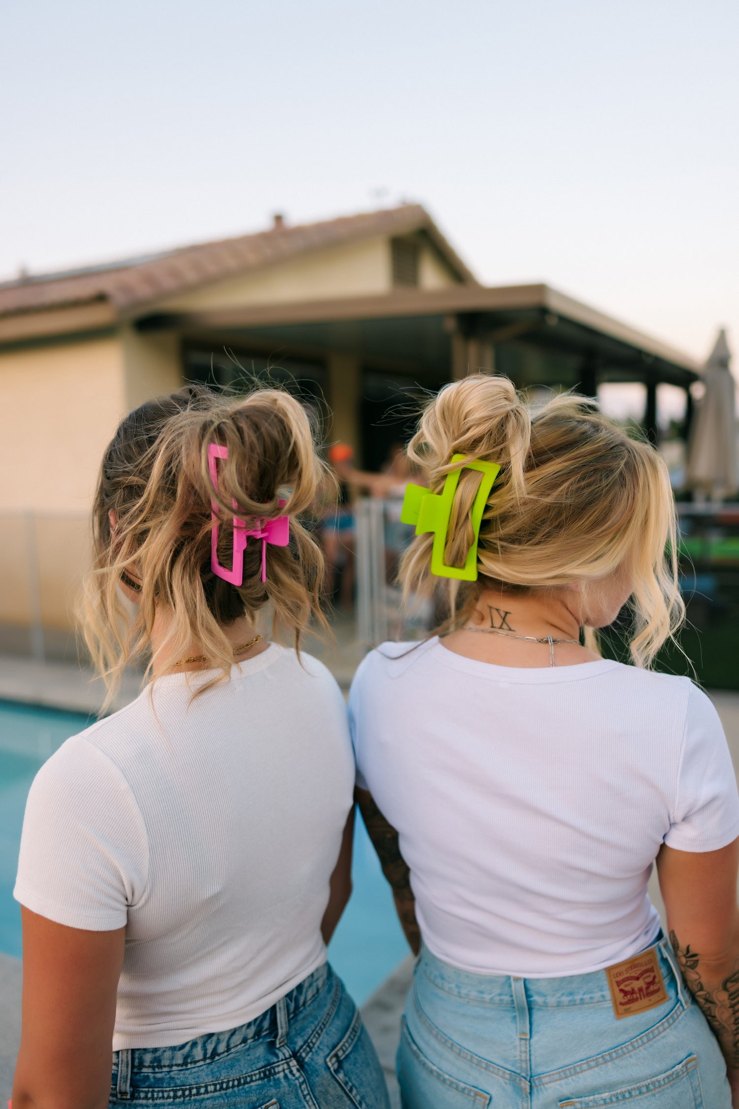 Two women with blonde hair in messy buns, secured with Mama X™ Brand Neon Green Claw Clip - XL and oversized clips, stand poolside in white shirts and blue jeans. Their thick hair is perfectly styled; a house and people blur in the background.