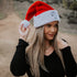 A woman with long blonde hair wears the Mama X™ Brand Mama X Santa Hat and a black top outdoors, smiling slightly as she holds the pom-pom. She also wears a gold heart necklace.