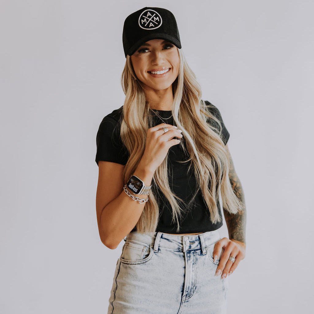A smiling woman with long blonde hair wears the Black Corduroy Mama X Trucker by Mama X™ Brand, a black t-shirt, and light-wash jeans. She holds a necklace and wears a smartwatch, standing against a plain light background.