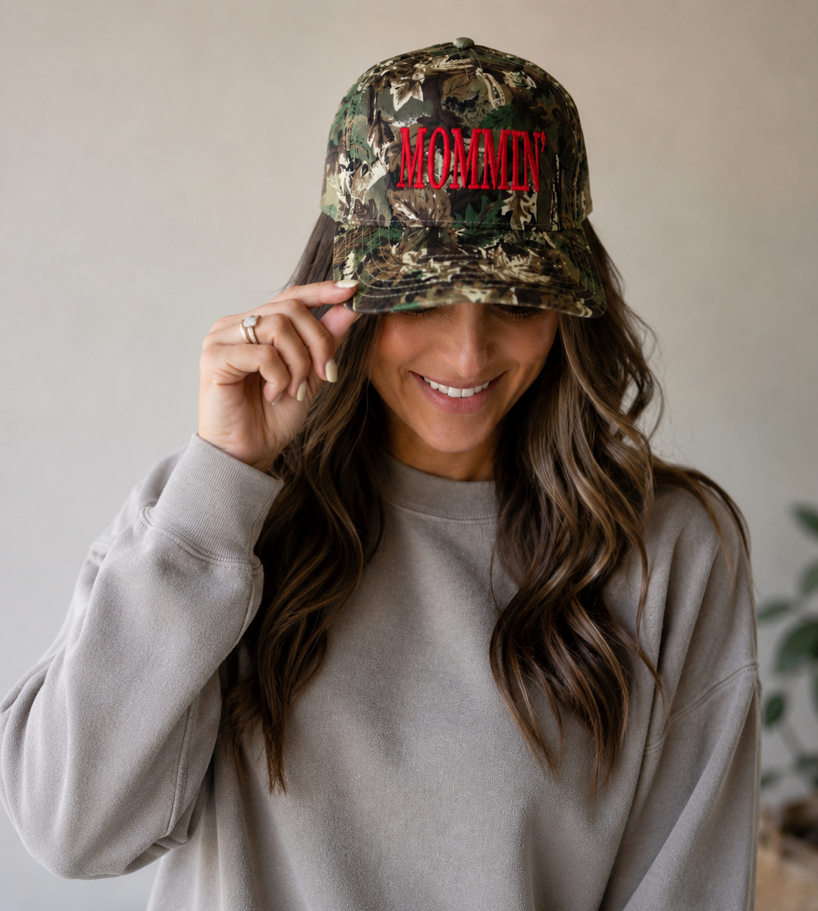 A woman with long wavy hair smiles, wearing the Mama X™ Brand "Mommin camo Red" baseball cap and a beige sweatshirt, holding the brim of her hat with one hand.