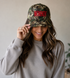 A woman with long wavy hair smiles, wearing the Mama X™ Brand "Mommin camo Red" baseball cap and a beige sweatshirt, holding the brim of her hat with one hand.