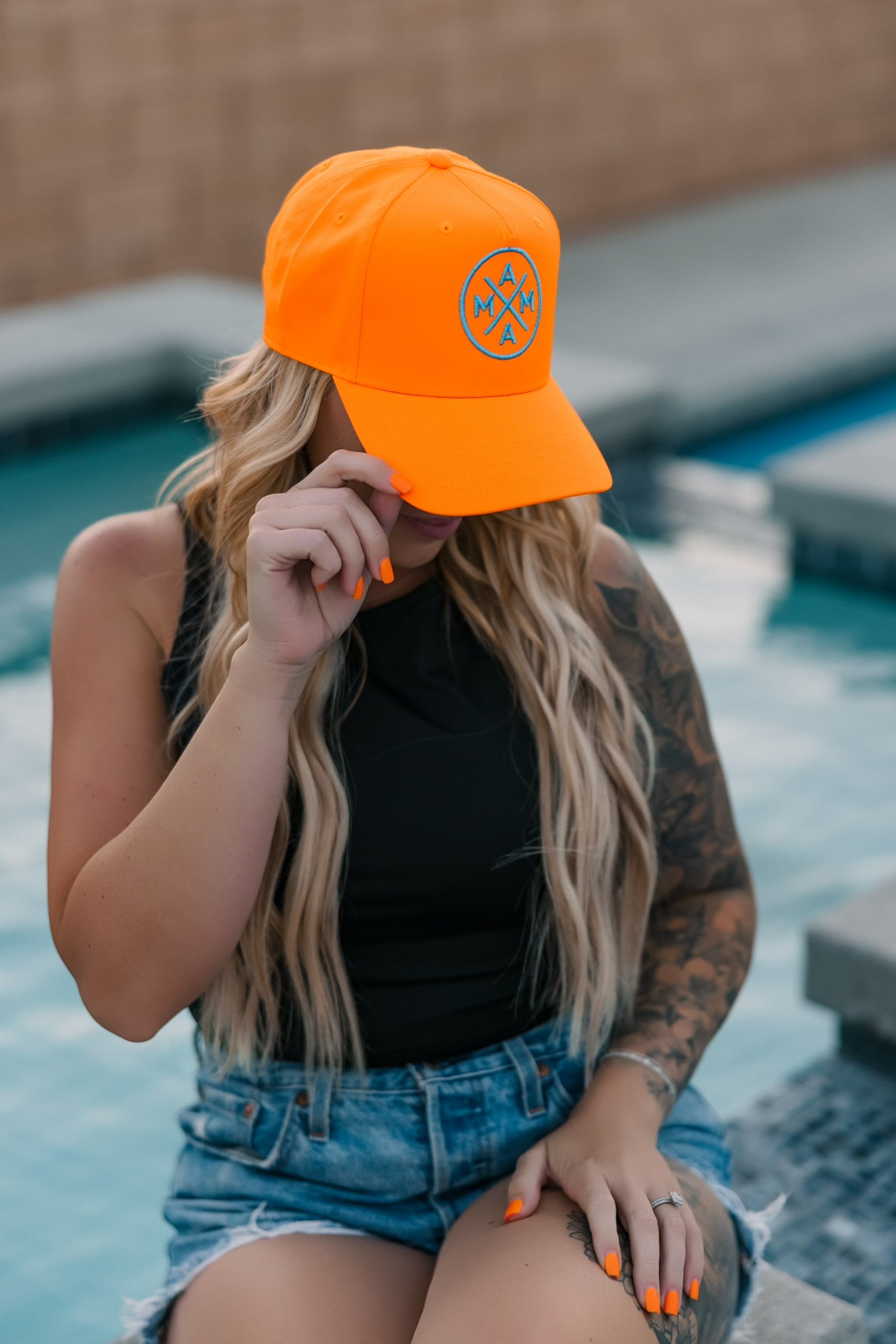 A person with long blonde hair, wearing the Neon Orange Mama X hat by Mama X™ Brand, a black sleeveless top, and denim shorts, sits poolside and covers part of their face with the cap. Their nails match in orange.