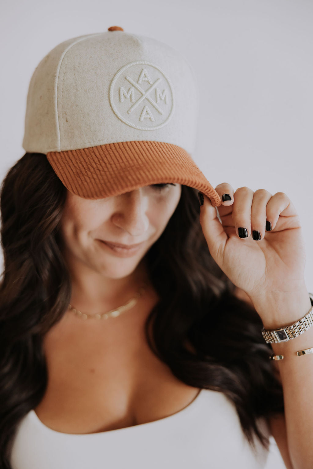 A woman with long, wavy dark hair wears Courtney’s Trucker Hat from Mama X™ Brand—a beige and brown felt cap with "MAMA" stitched on the front. She holds the corduroy brim, looks down, and pairs it with a white top and bracelets.