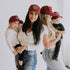 Two women in Mama X™ Brand Maroon Suede Mama X Trucker hats and white tops hold two children, all wearing matching red caps. The smiling group poses together against a white background.