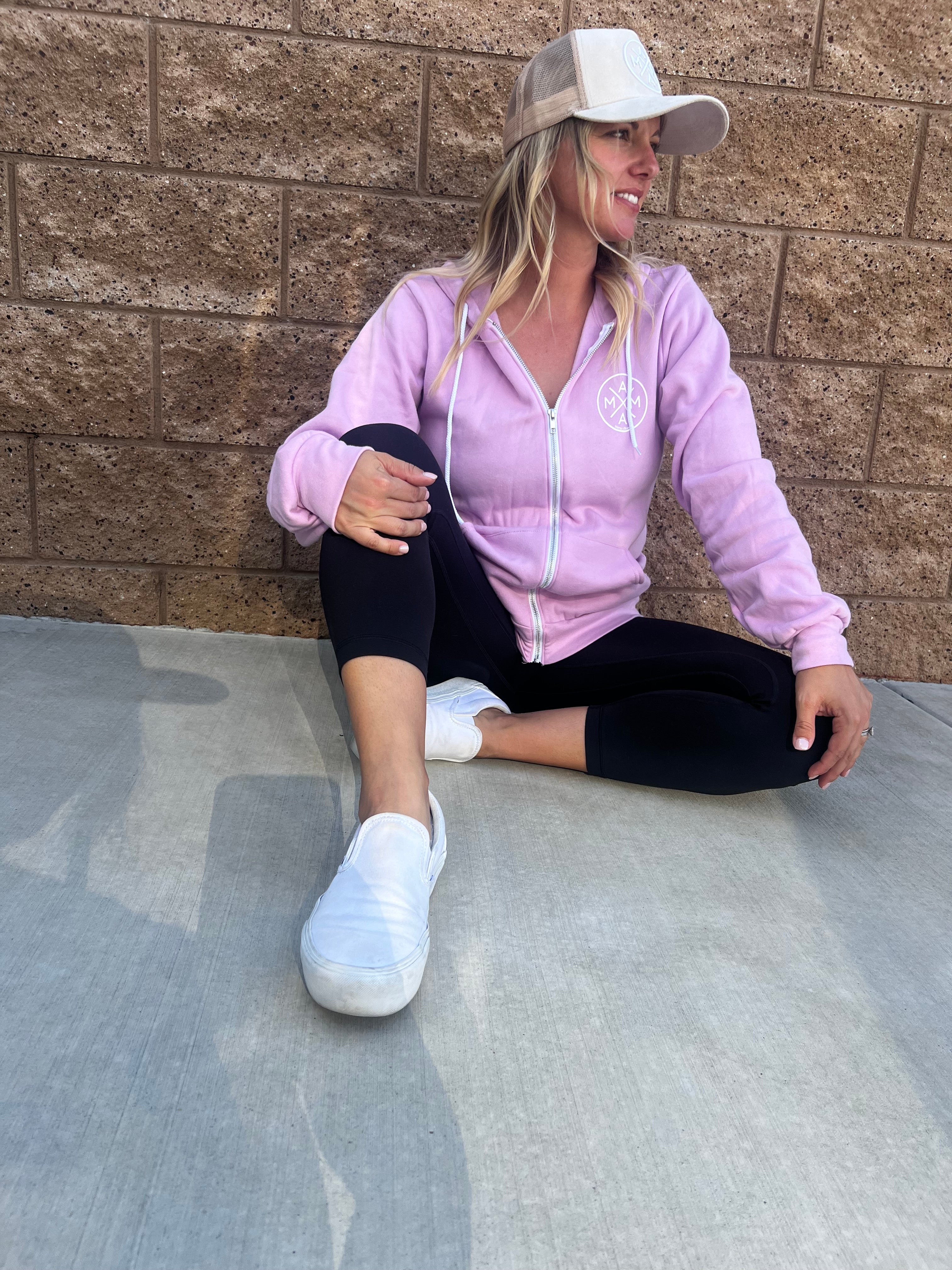 A person with long hair sits on the ground against a brick wall, wearing a beige cap, a lavender Mama X™ Zip Up Sweatshirt from Mama X™ Brand made from premium ring-spun cotton, black leggings, and white slip-on shoes. They are looking to the side and smiling with one leg bent and the other stretched out.