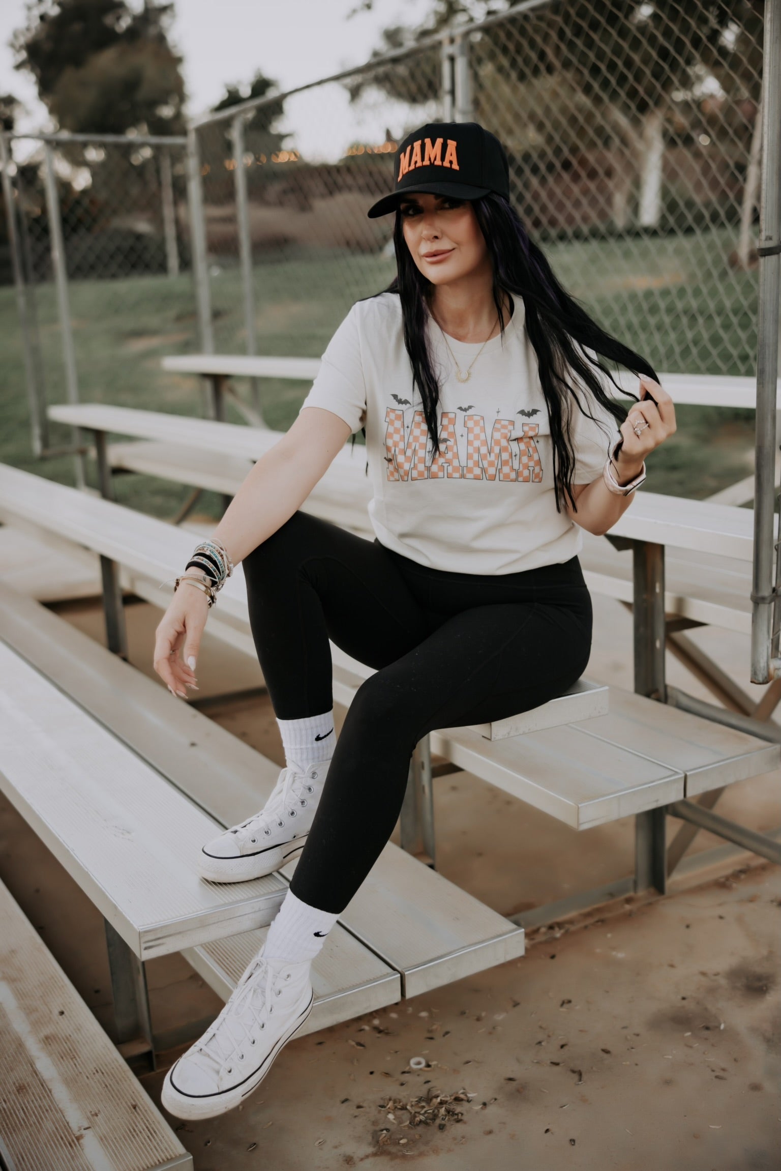 A woman sits on metal bleachers outdoors, smiling and wearing a Mama Halloween Tshirt by Mama X™ Brand, paired with black leggings, white high-top sneakers, and a black "MAMA" cap as she holds her long dark hair.