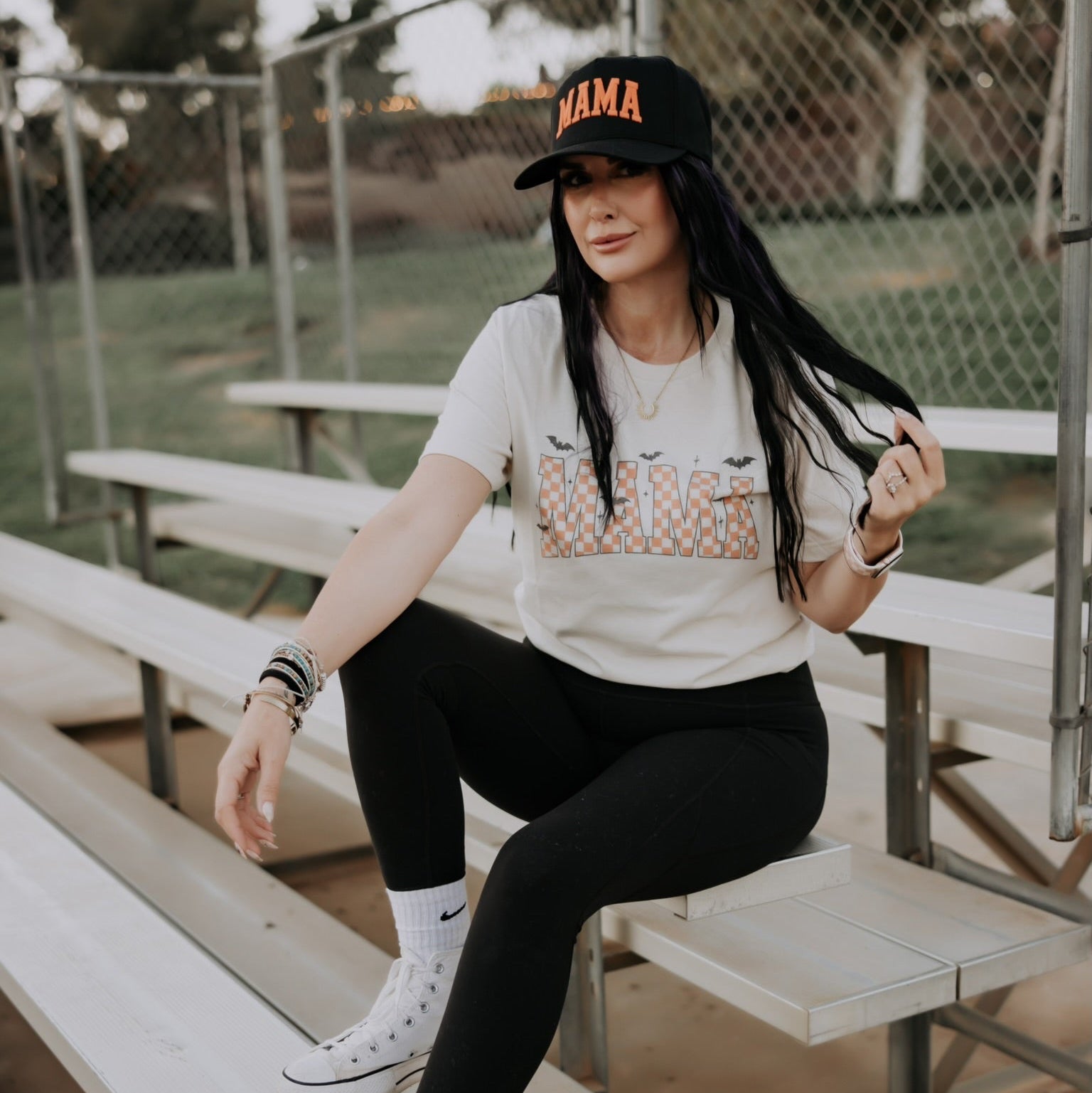 A woman sits on metal bleachers outdoors, smiling and wearing a Mama Halloween Tshirt by Mama X™ Brand, paired with black leggings, white high-top sneakers, and a black "MAMA" cap as she holds her long dark hair.