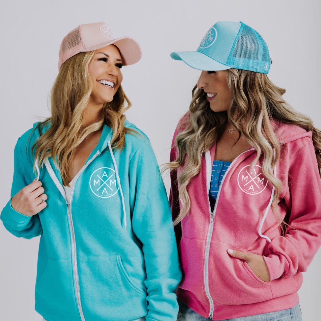 Two smiling women are facing each other, both donning Mama X™ Zip Up Sweatshirts—one in turquoise and the other in pink. Each unisex hoodie features a circular logo on the chest and a cap that matches its color. The backdrop is a plain, light shade.