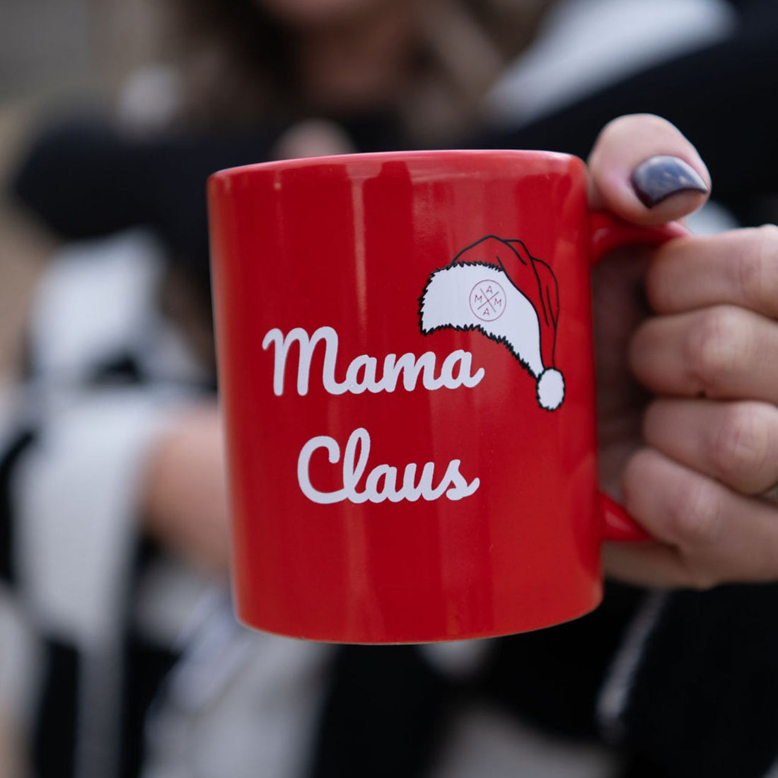 A person holds the Mama Claus Mug by Mama X™ Brand—a red mug with "Mama Claus" in white letters and a Santa hat graphic. In the blurred background, two people are smiling.