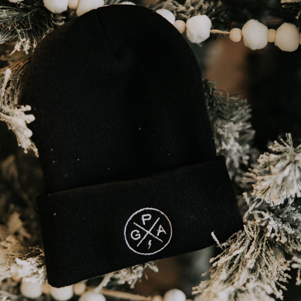 The GPA Cuffed Beanie by Mama X™ Brand—a black beanie with a white circular “GPA” logo—hangs on a festive, snow-dusted evergreen branch, making a soft, warm Christmas gift for Grandpa.
