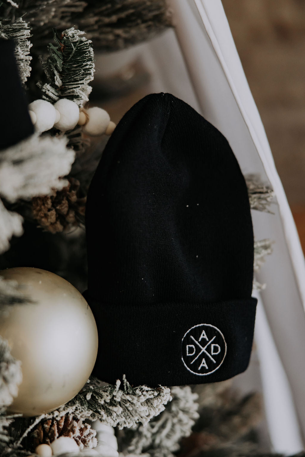 The Dada Cuffed Beanie by Mama X™ Brand, featuring a circular white "DADA" logo, is shown on a snow-dusted Christmas tree with festive accents—making it the perfect holiday gift for Dada.