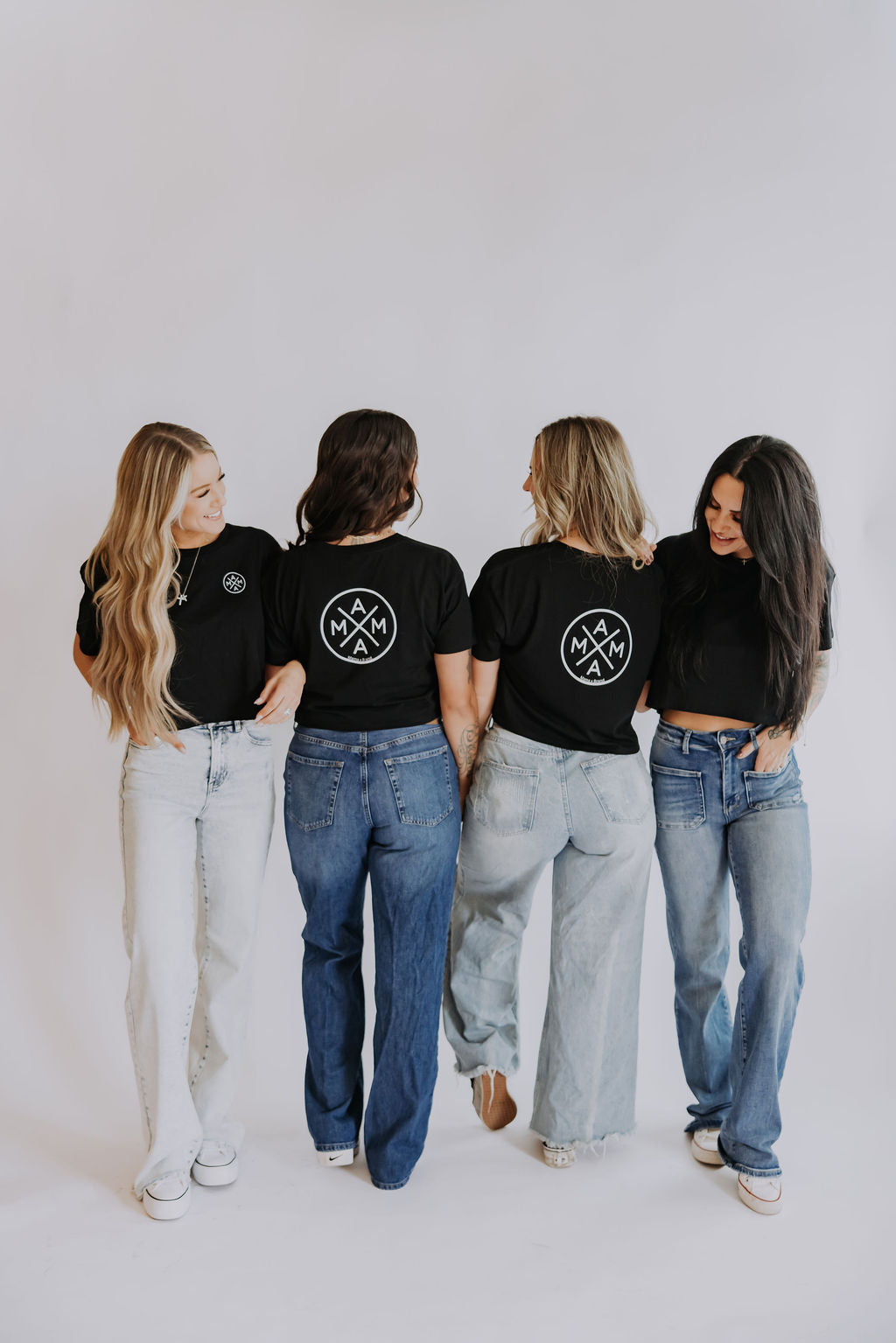 Four women pose against a white background in relaxed fit blue jeans and Mama X™ Brand "Mama X crop top" black shirts. Three show the circular "MAMA" logo on the back, while one smiles at the group in her comfy combed cotton top.