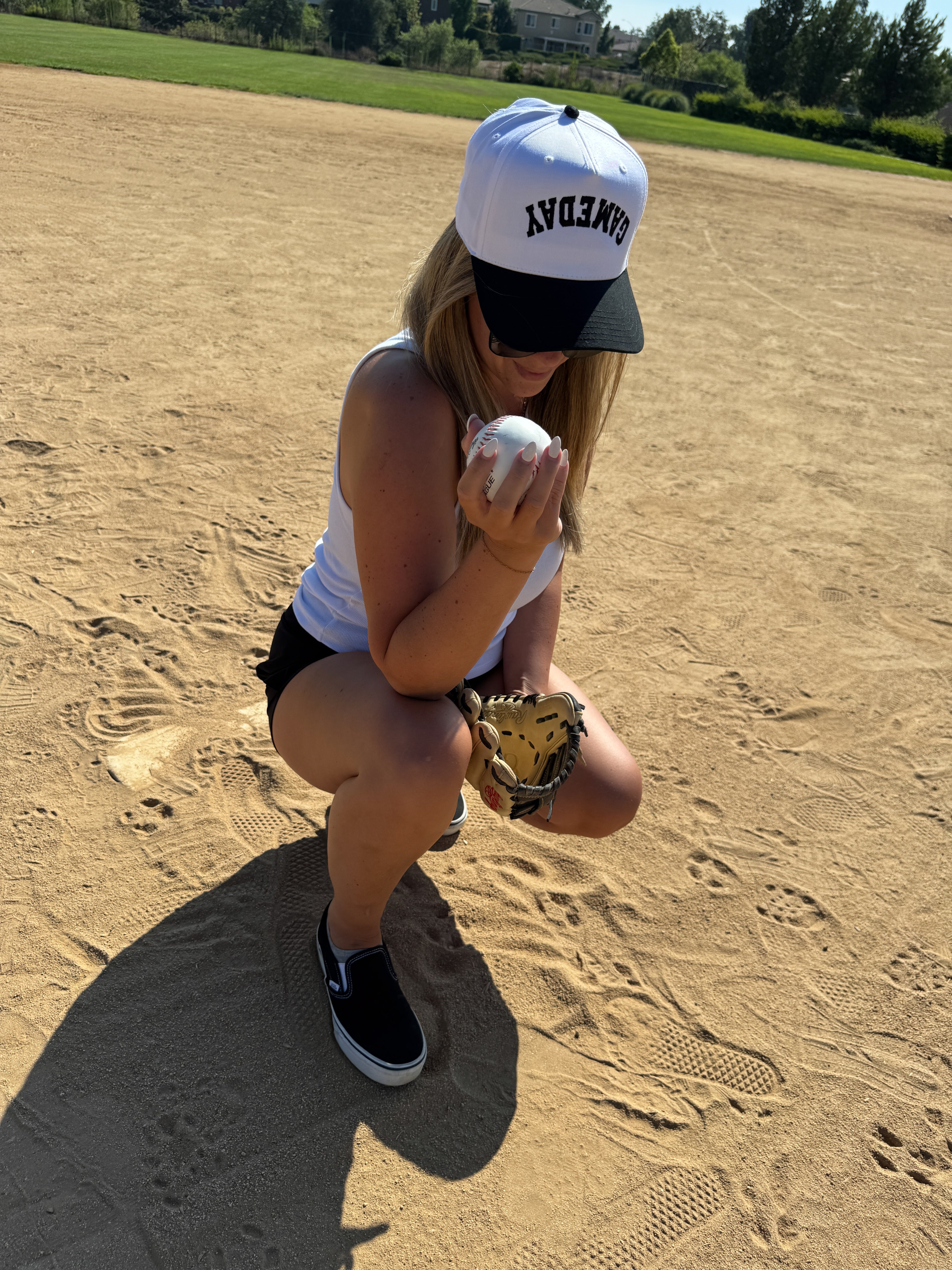 A person in a white tank top and black shorts kneels on a sunny baseball field, wearing the Mama X™ Brand Game Day Trucker hat, holding a baseball in one hand and a glove in the other.