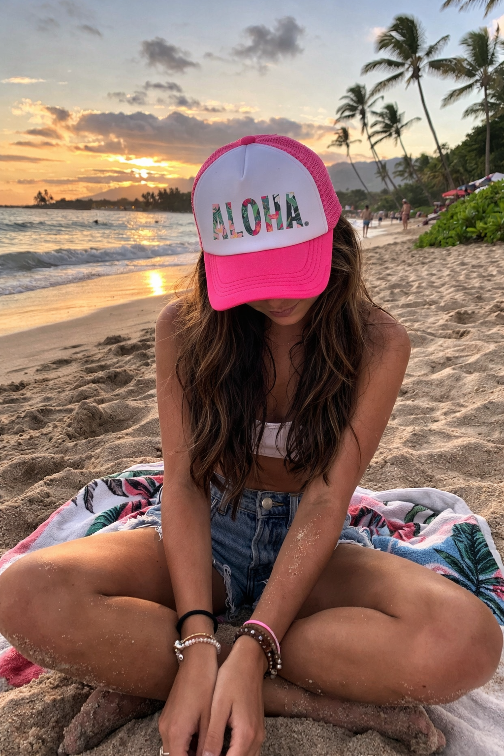 At sunset on a sandy beach, a woman in a Mama X™ Brand Aloha Trucker Hat, white top, and denim shorts sits cross-legged among palm trees by the ocean, her face partially hidden as she gazes down.