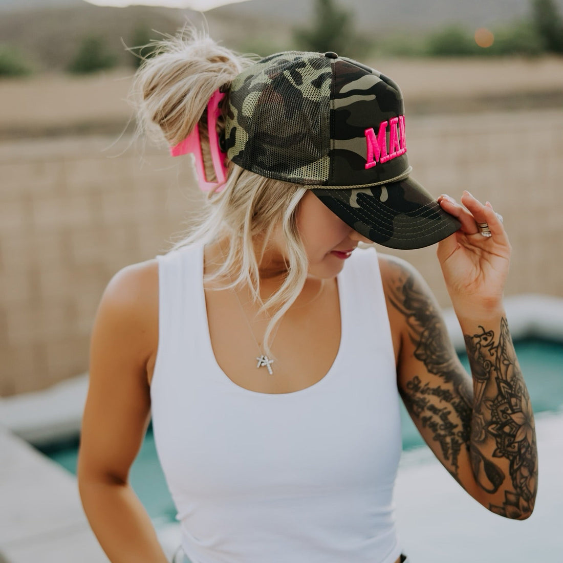 A woman with a tattooed arm wears the Mama X™ Brand Foam Neon Pink Mama Trucker, a white tank top, and denim shorts while standing poolside with mountains and a fence in the background.