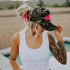 A woman with a tattooed arm wears the Mama X™ Brand Foam Neon Pink Mama Trucker, a white tank top, and denim shorts while standing poolside with mountains and a fence in the background.