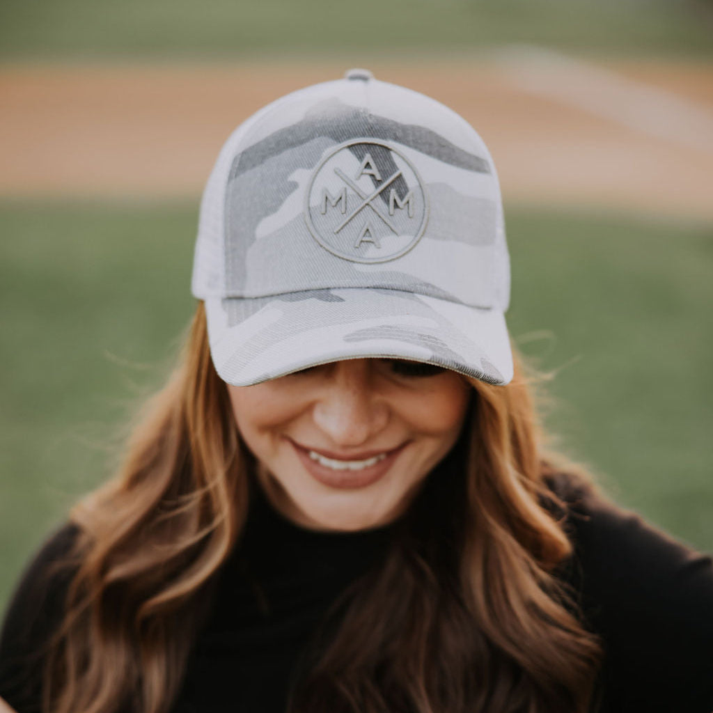Wearing the White Camo Mama X™ Trucker Hat by Mama X™ Brand, featuring "MAMA" on the front, a person showcases their flowing brown hair over a black shirt. The adjustable snapback ensures mom pride shines against a blurred baseball field backdrop.