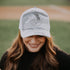Wearing the White Camo Mama X™ Trucker Hat by Mama X™ Brand, featuring "MAMA" on the front, a person showcases their flowing brown hair over a black shirt. The adjustable snapback ensures mom pride shines against a blurred baseball field backdrop.