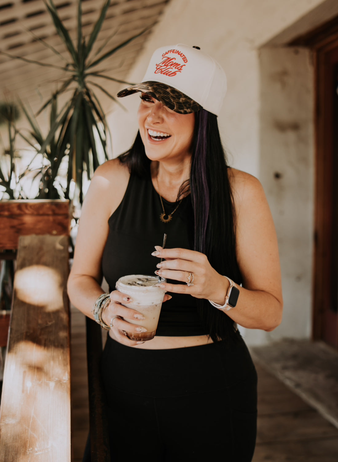 A woman with long dark hair smiles while holding a cold drink, wearing the Mama X™ Brand Caffeine Club Trucker cap. She stands indoors near a plant and wooden railing, dressed in a black sleeveless top and black pants.