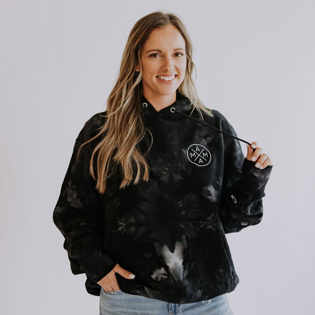 A woman with long blonde hair smiles while wearing a Unisex Champion tie-dye hoodie by Mama X™ Brand and ripped light blue jeans, holding one hoodie string with her right hand against a plain white background.