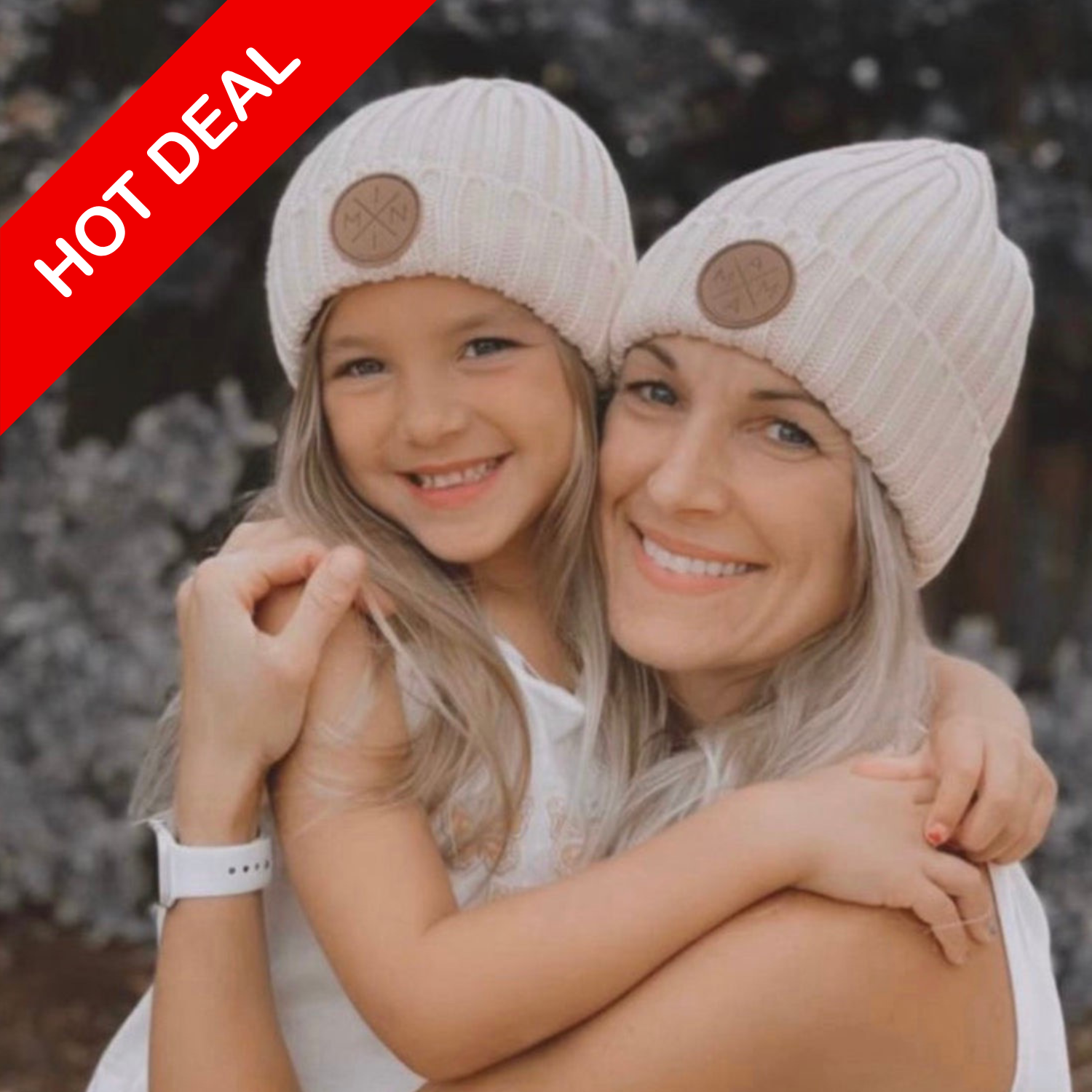 A woman and young girl smile warmly, both sporting matching Mini X knit beanies with brown leather patches from Mama X™ Brand. The woman gently holds the girl in her arms as a 