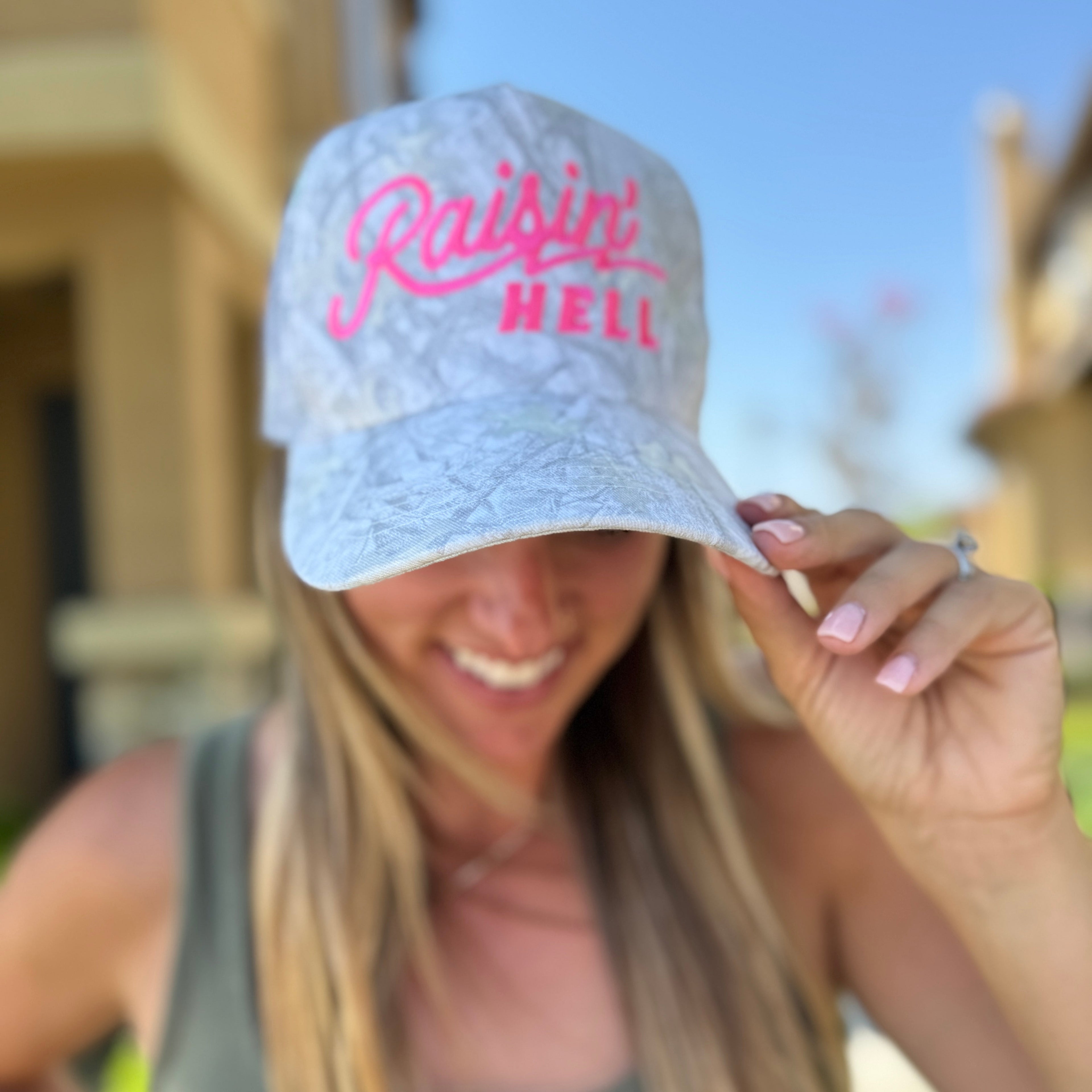 A smiling woman outdoors in a tank top shows off the Mama X™ Brand “Raisin’ Hell” Camo Trucker cap, playfully embodying motherhood spirit. The blurred background features sunlit buildings and greenery.