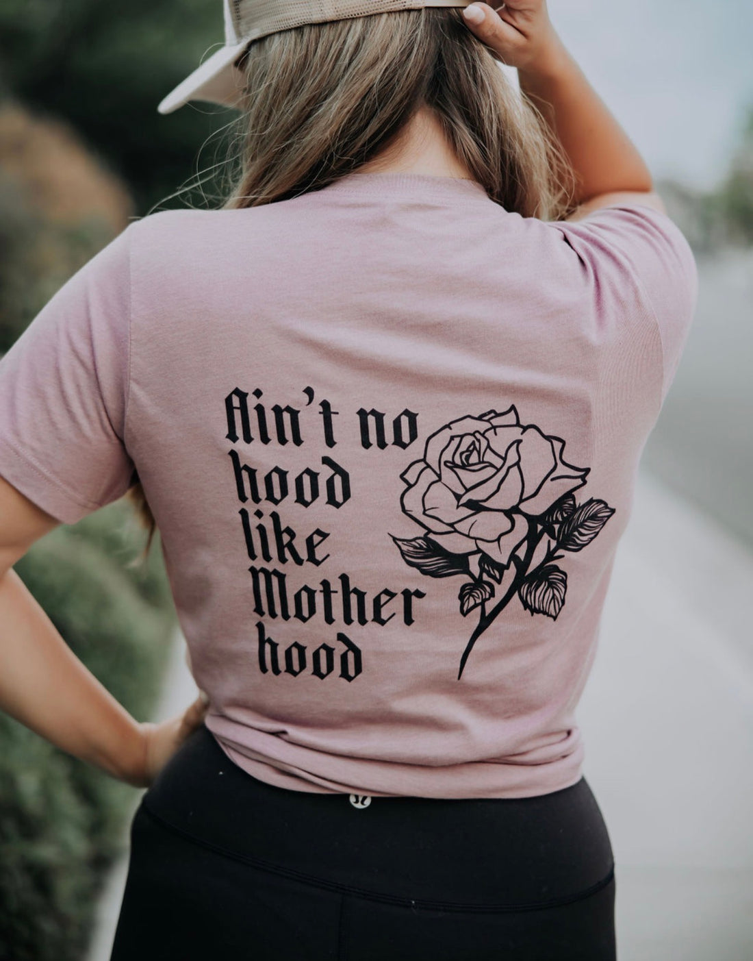 Wearing the Mama X™ Brand Motherhood Tshirt in soft light pink with black “Ain’t no hood like Motherhood” text and a rose illustration on the back, this person pairs the relaxed fit tee with a beige cap, hand resting on their hip.