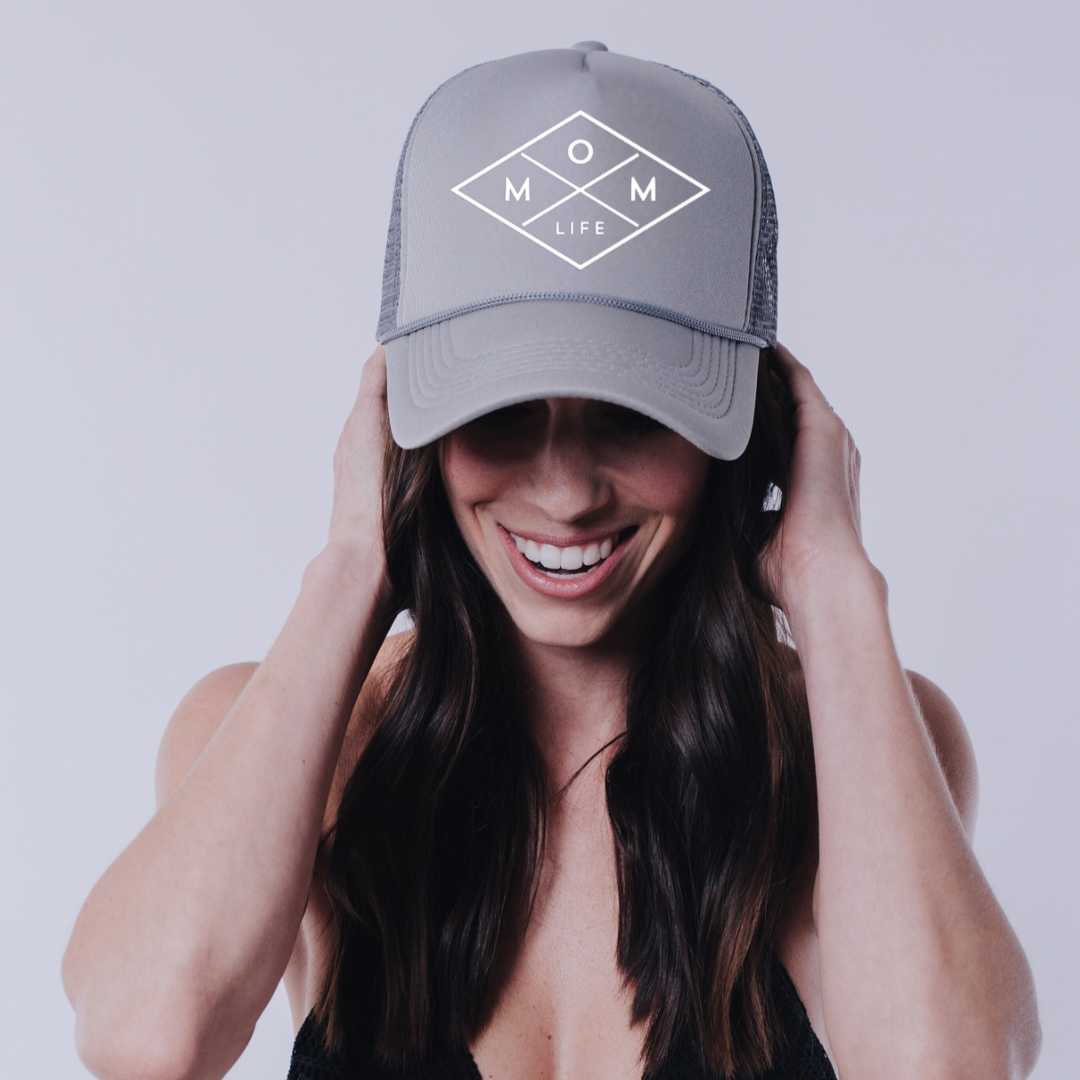 A woman with long dark hair is smiling and looking down while wearing a gray Mom Life Foam Hat from Mama X™ Brand, featuring a diamond-shaped design on the front. She holds the sides of the hat with both hands against a plain white background.