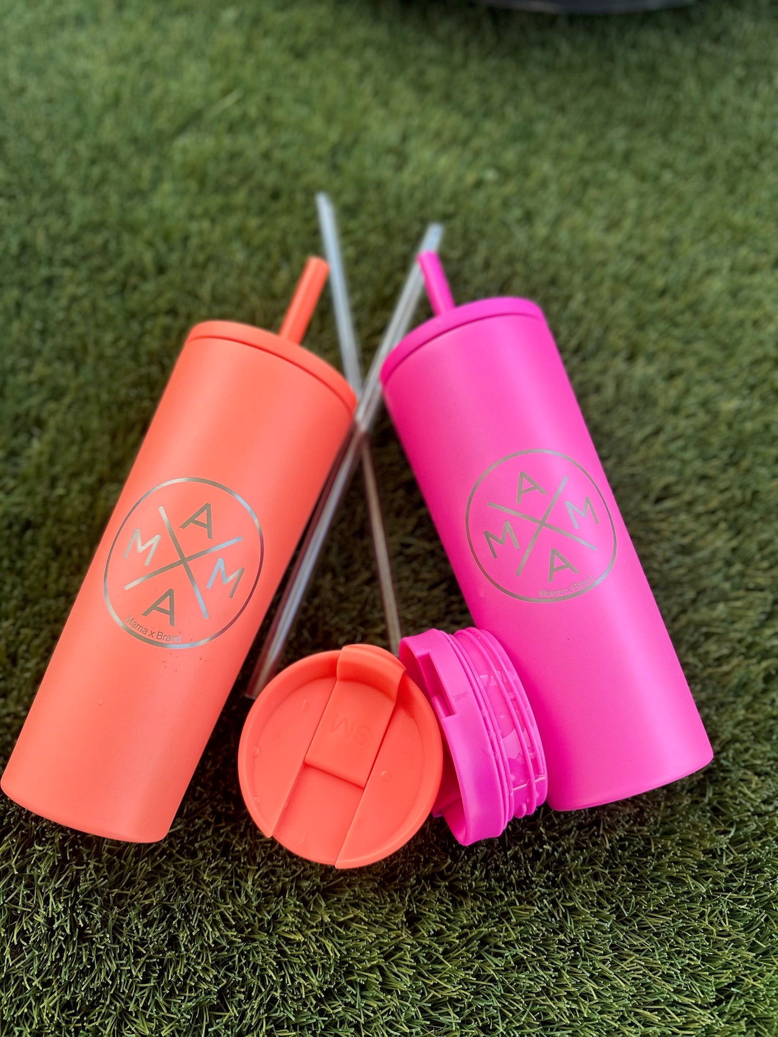 Two Mama X™ Brand Coral Tumblers lie on green grass with their lids and straws detached and placed beside them.