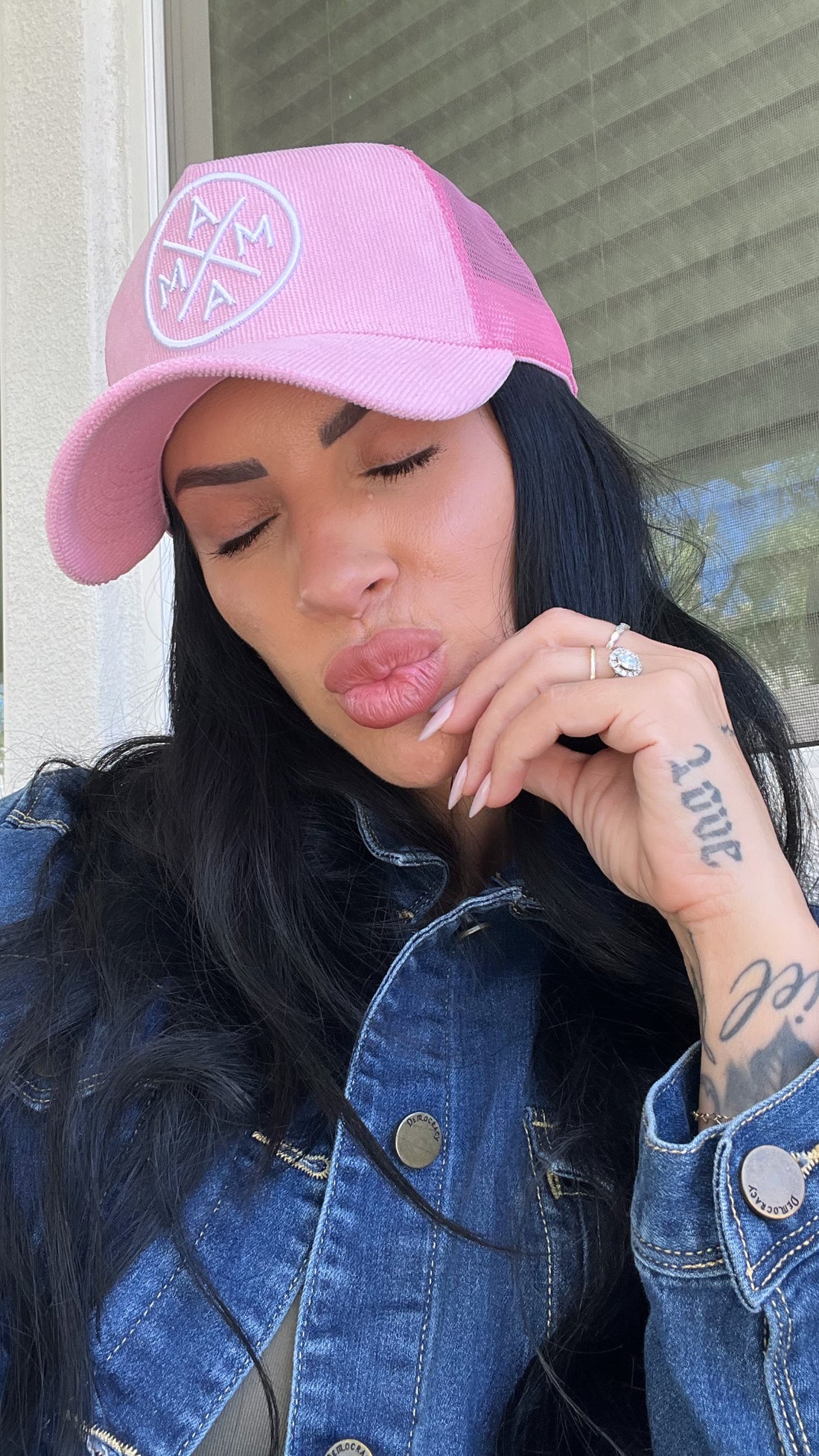 A woman with long dark hair, wearing a Pink Corduroy Mama X Trucker hat by Mama X™ Brand and a denim jacket, closes her eyes and puckers her lips while showing off a ring and revealing a “LOVE” tattoo on her hand.