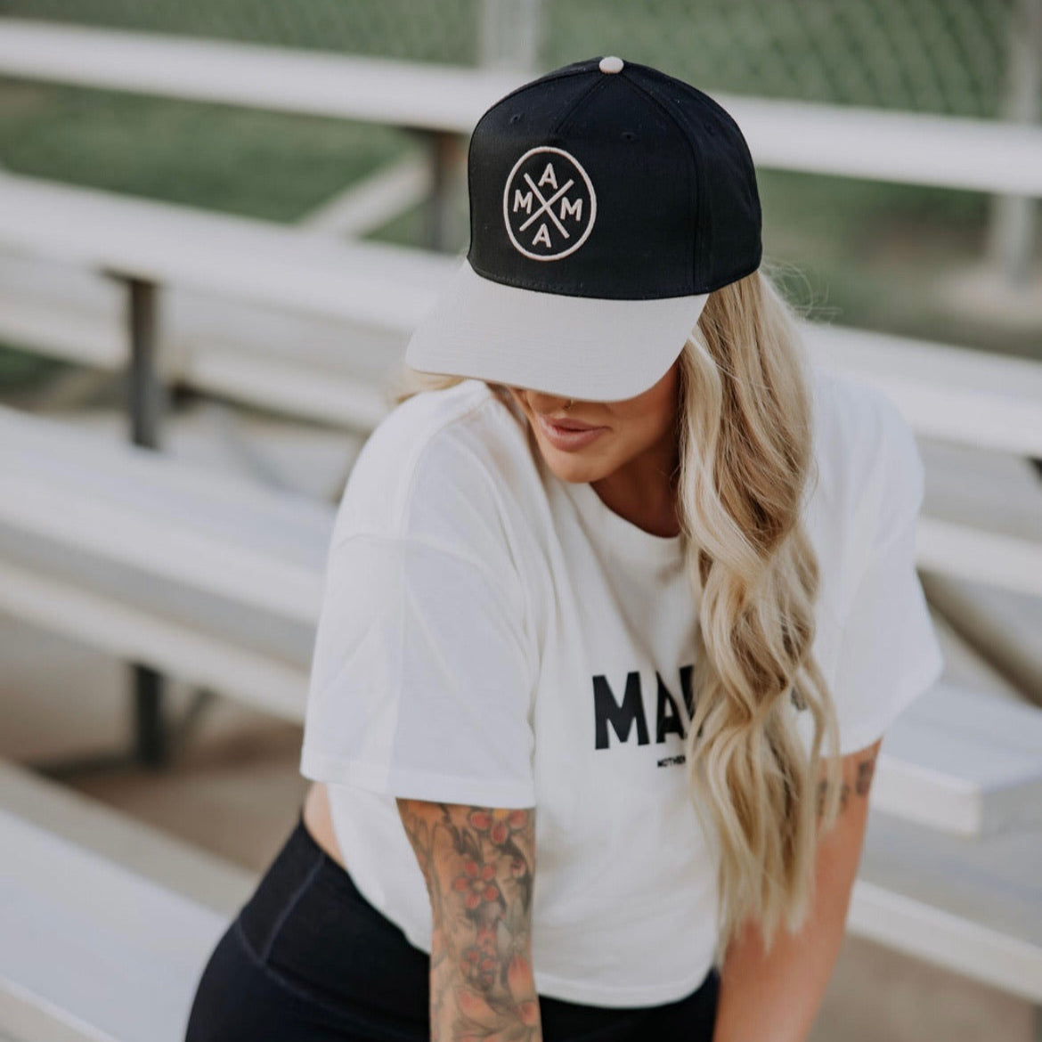 A woman with long blonde hair and tattooed arms sits on bleachers wearing the Mama X™ Trucker hat by Mama X™ Brand and a white tee, looking downward. A chain-link fence and grassy field are in the background.
