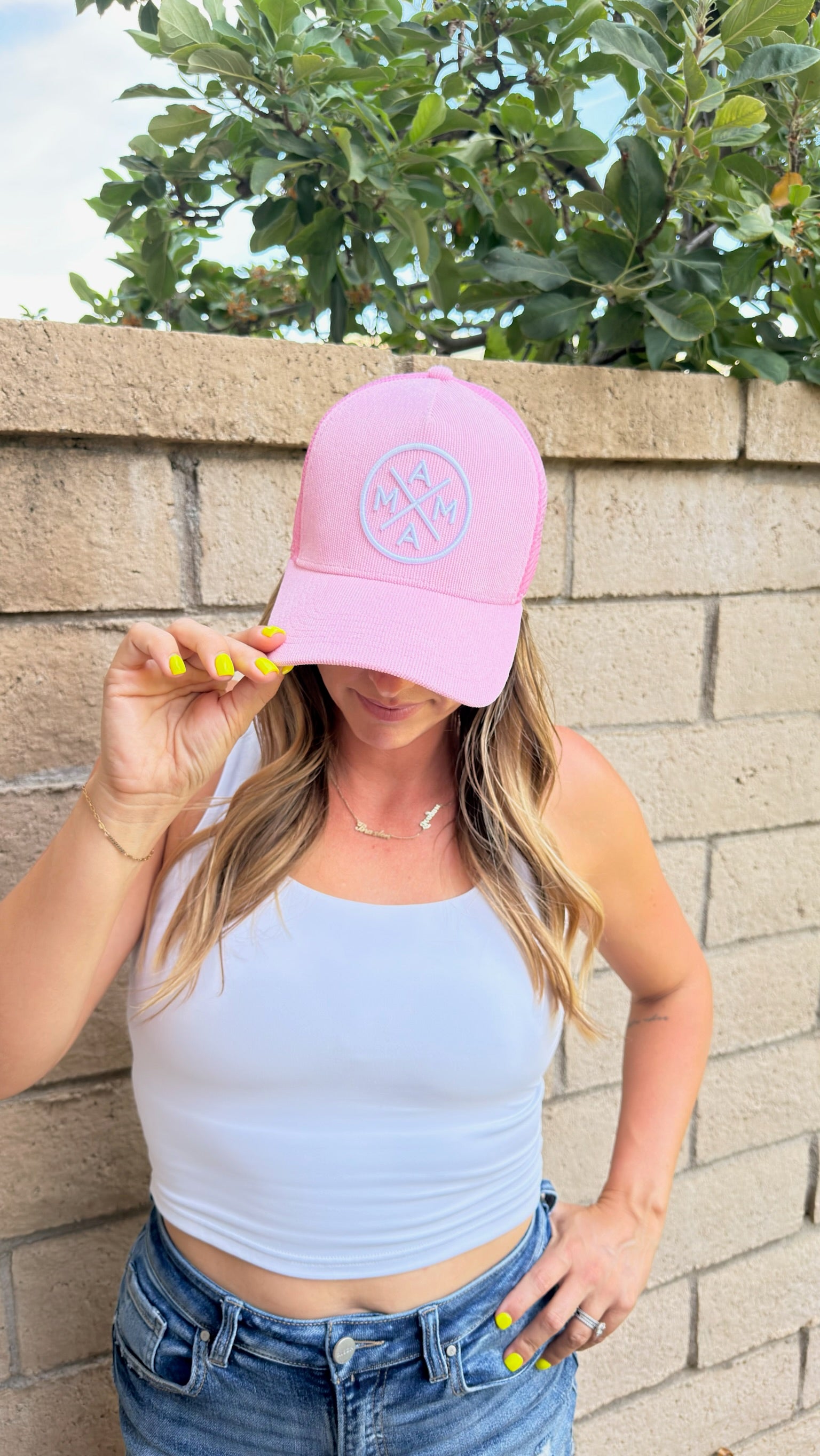 A woman wearing a white tank top and blue jeans stands in front of a brick wall, holding the brim of her Mama X™ Brand Pink Corduroy Mama X Trucker hat with one hand. The hat features “MAMA” on the front. Her nails are painted yellow, and green leaves hang above the wall.