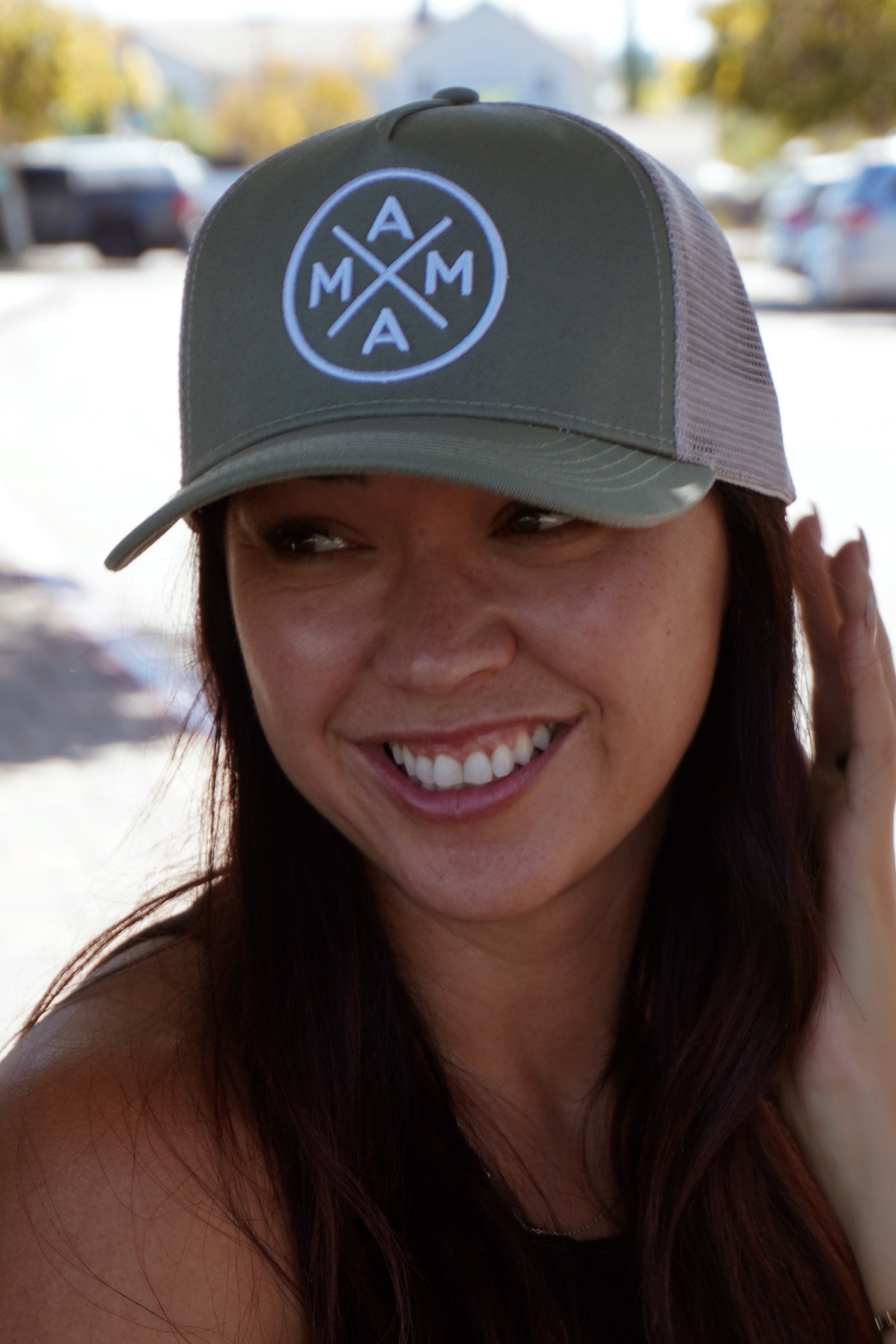 A woman with long dark hair sits outdoors on a sunny day wearing the Mama X™ Brand Premium Canvas Trucker Hat in sage and a black tank top.