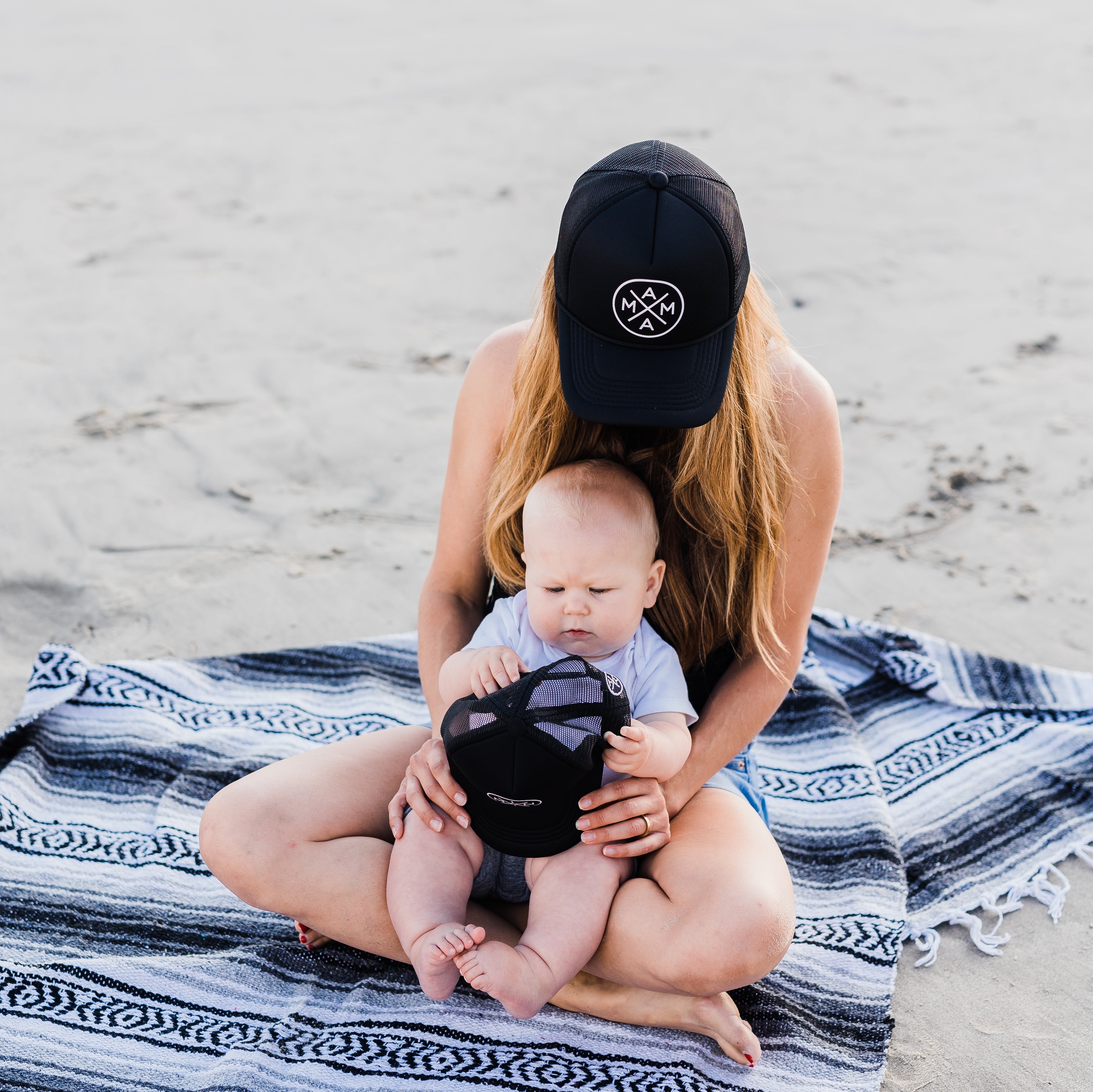 A person with long hair, wearing a best-seller Mama X™ Foam Trucker Hat from Mama X™ Brand, sits on a striped blanket at the beach holding a baby. The baby, also wearing a smaller version of the same high-quality 5-panel cap, is focused on the hat. Sand and ocean waves are visible in the background.