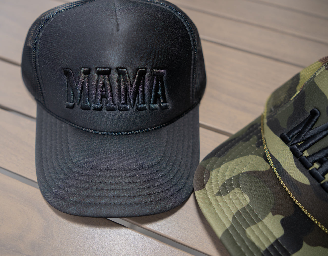 A Black out foam Trucker hat from Mama X™ Brand sits next to a camouflage-patterned hat on a wooden surface.