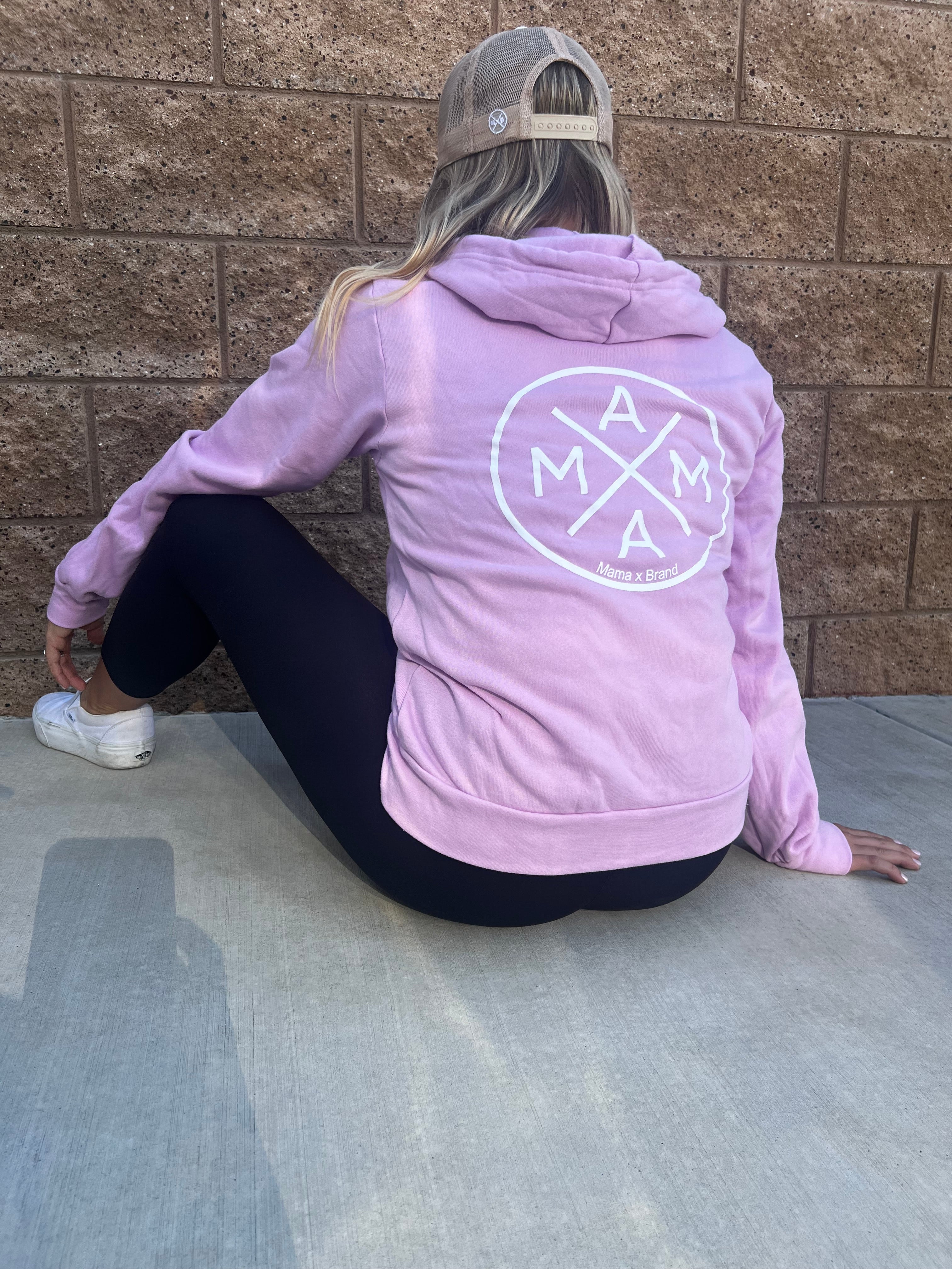 A person wearing a lavender Mama X™ Zip Up Sweatshirt made of premium ring-spun cotton with a circular white logo on the back and black leggings is sitting on a concrete surface against a brown brick wall. They are also wearing a beige baseball cap and white sneakers. The person is sitting with one arm resting on the ground.