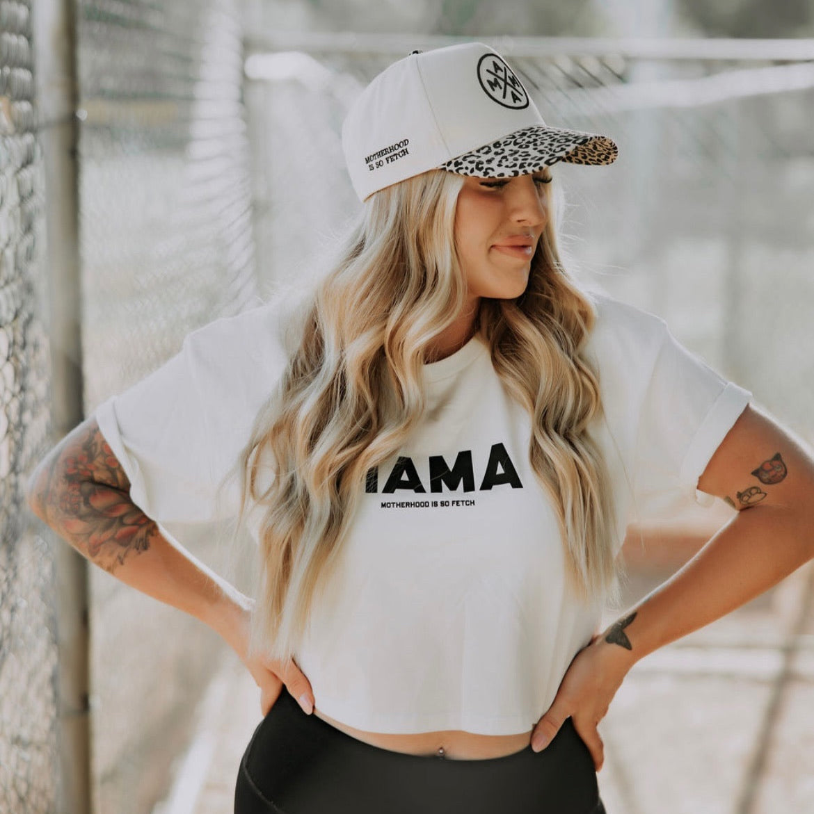 A woman with long wavy blonde hair wears the Mama X + Motherhood Is So Fetch Trucker by Mama X™ Brand and a "MAMA" tee, smiling outdoors near a chain-link fence—ready for an exclusive ambassador collaboration drop.