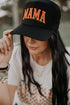 A woman with long dark hair, wearing a white shirt and jewelry, touches the brim of a Mama X™ Brand Black/Orange Mama Trucker hat that partially hides her face.