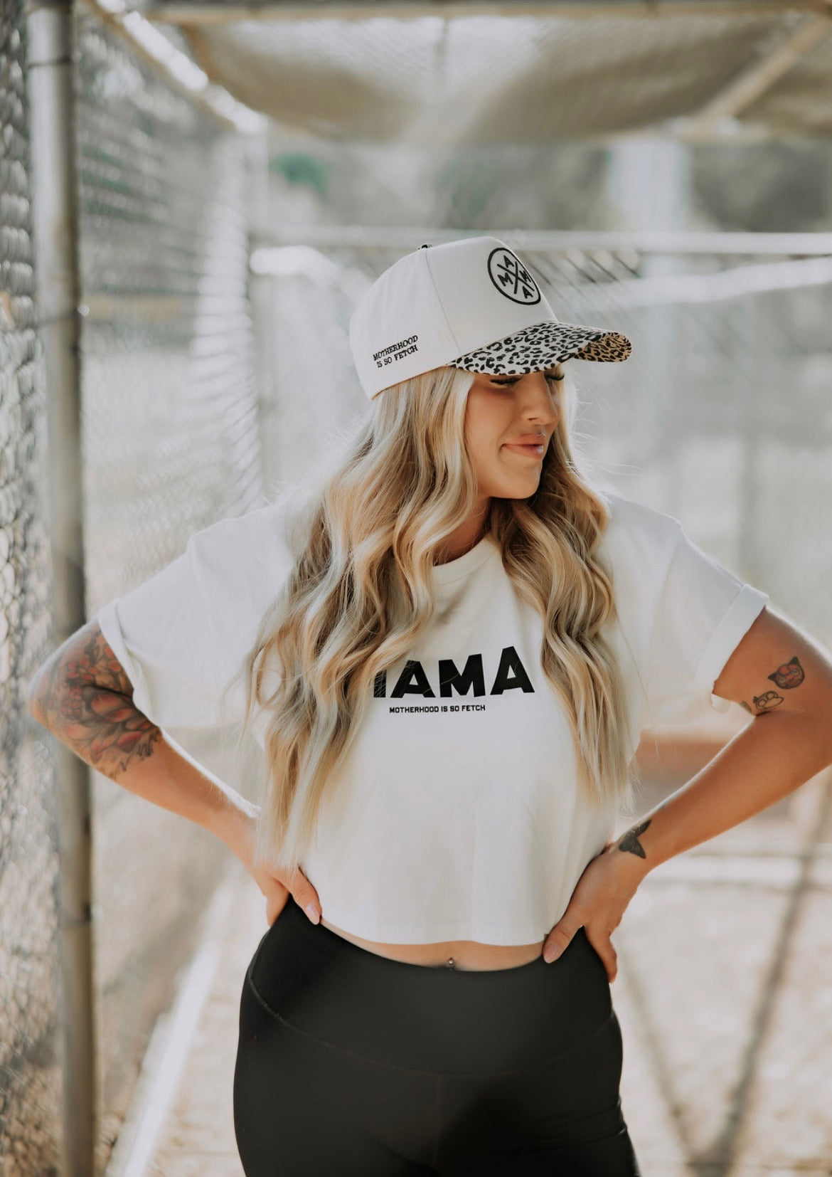 A woman with long blonde hair stands in a sunlit walkway, hands on hips, wearing the Mama X™ Brand "Motherhood Is So Fetch" crop top and black leggings. She has tattoos on both arms and looks to the side.
